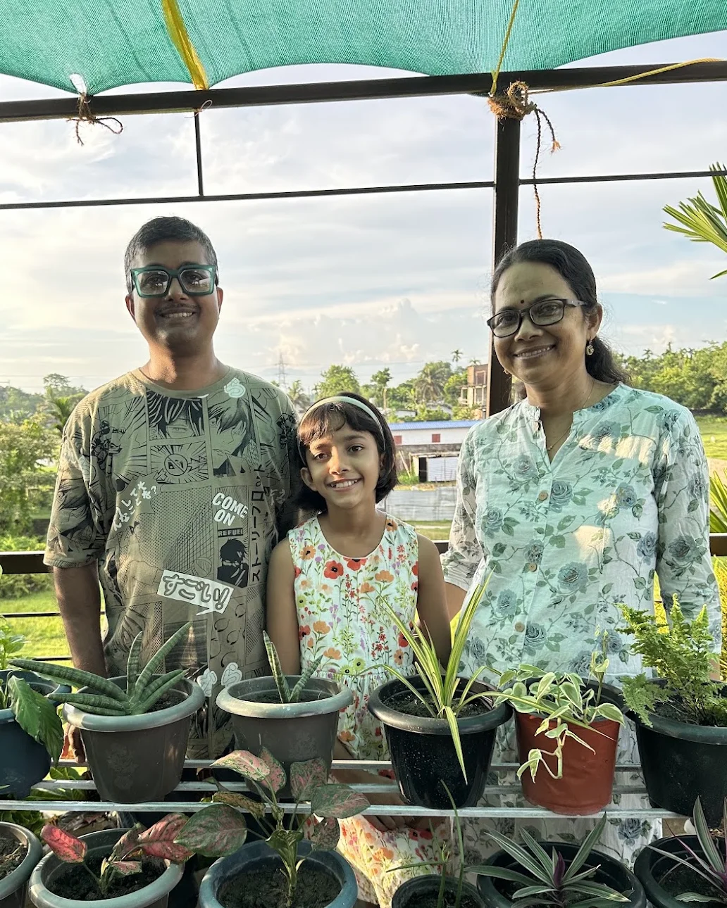 The three of us standing behind tiered shelves of potted plants under a green shade net.